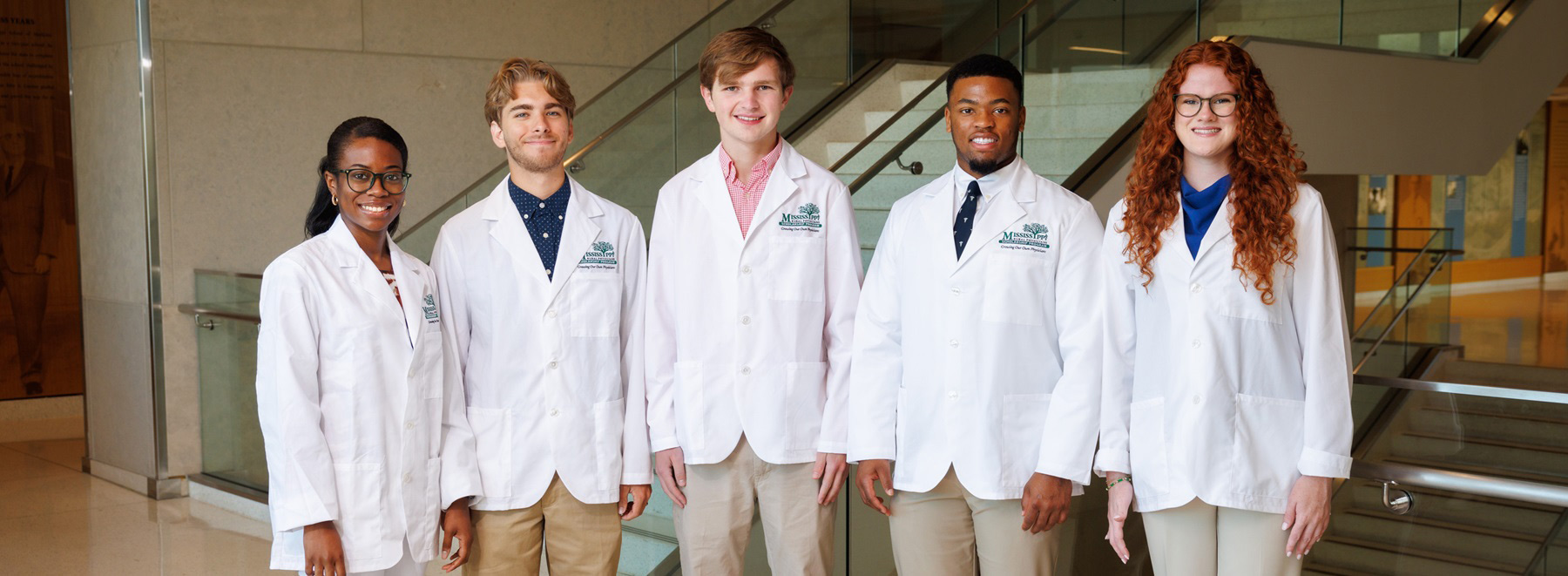 Five medical students wearing white lab coats stand side by side inside a modern academic building near a staircase.